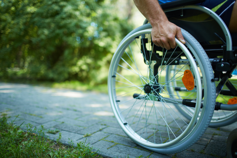 Primo piano di una mano maschile su ruota della sedia a rotelle durante la passeggiata nel parco - © Pressmaster/Shutterstock