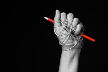 Hand with red pencil tied with rope, depicting the idea of freedom of the press or freedom of expression on a dark background © siam.pukkato/Shutterstock