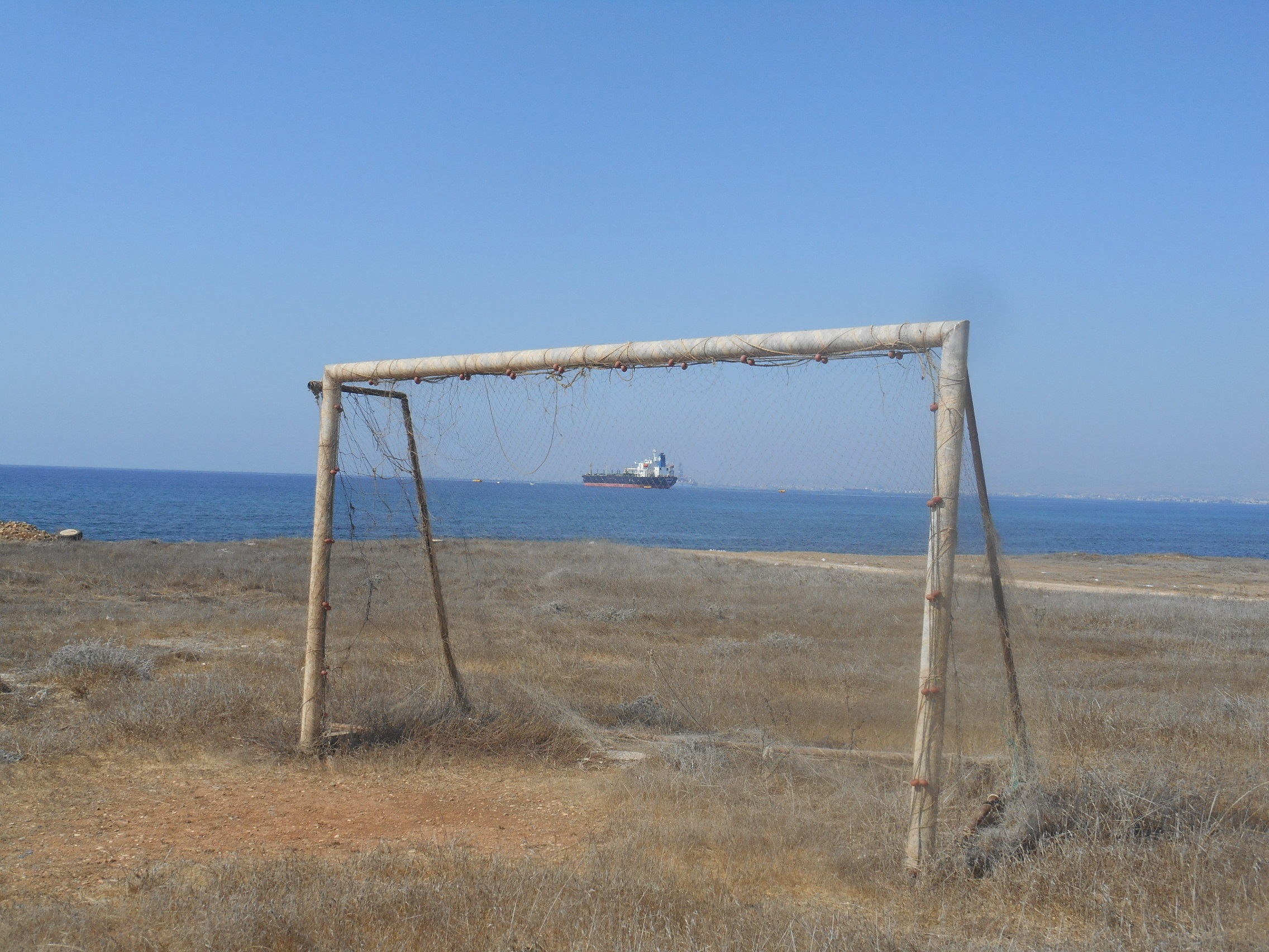 Strada statale B3, Cipro, omaggio a Luigi Ghirri. Un campetto da calcio deserto e bruciato dal sole, una porta attrezzata con una vecchia rete da pesca, un Egeo indaco e una petroliera alla fonda, davanti alla Centrale Termoelettrica di Dhekelia, 15 chilometri a nord di Larnaca - Fabio Fiori