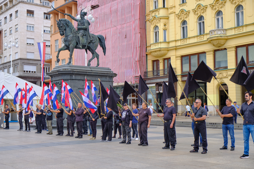Zagreb, Croatia, June 23, 2019 : Right wing political rally in city main square, featuring man dressed in black waving Croatian, Black and Anti EU flags (© meandering images/Shutterstock) Zagreb, Croatia, June 23, 2019 : Right wing political rally in city main square, featuring man dressed in black waving Croatian, Black and Anti EU flags (© meandering images/Shutterstock)