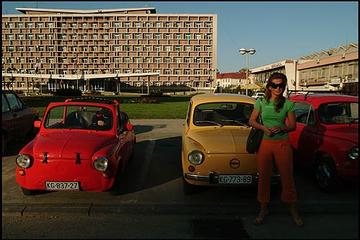 Sullo sfondo un edificio socialista, in primo piano una ragazza ed alcune macchine Zastava