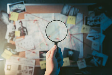 Detective mano che tiene una lente di ingrandimento di fronte a una tavola con prove, foto della scena del crimine e mappa (© Fer Gregory/Shutterstock)